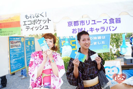 「京都市リユース食器お試しキャラバン」の幕の前で、浴衣の二人が配りものをしている