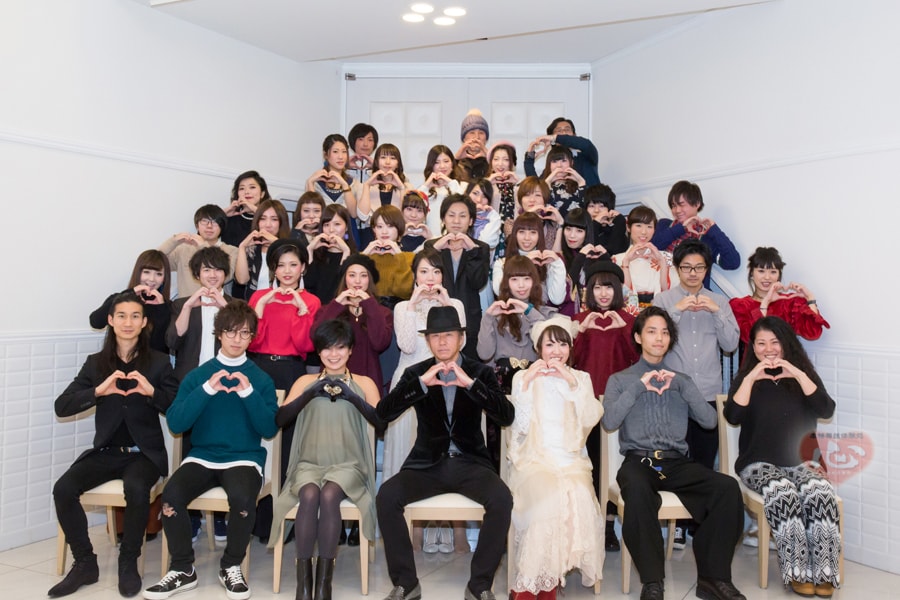 白い部屋で、三十人ほどが指でハートをつくって並ぶ