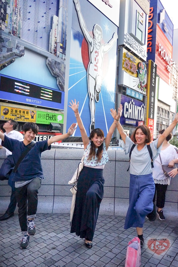 大きな看板の並ぶ道で、三人が飛びはねている