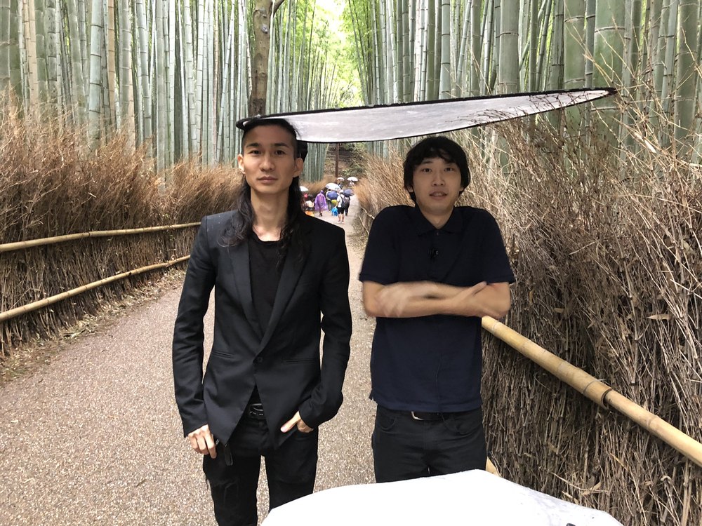 竹林の道で、黒い服の二人が並んで立つ