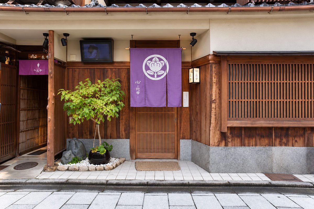 祇園店「心-花雫-」の外観。町家の格子戸に、白い紋の入った紅い暖簾が掛かっている