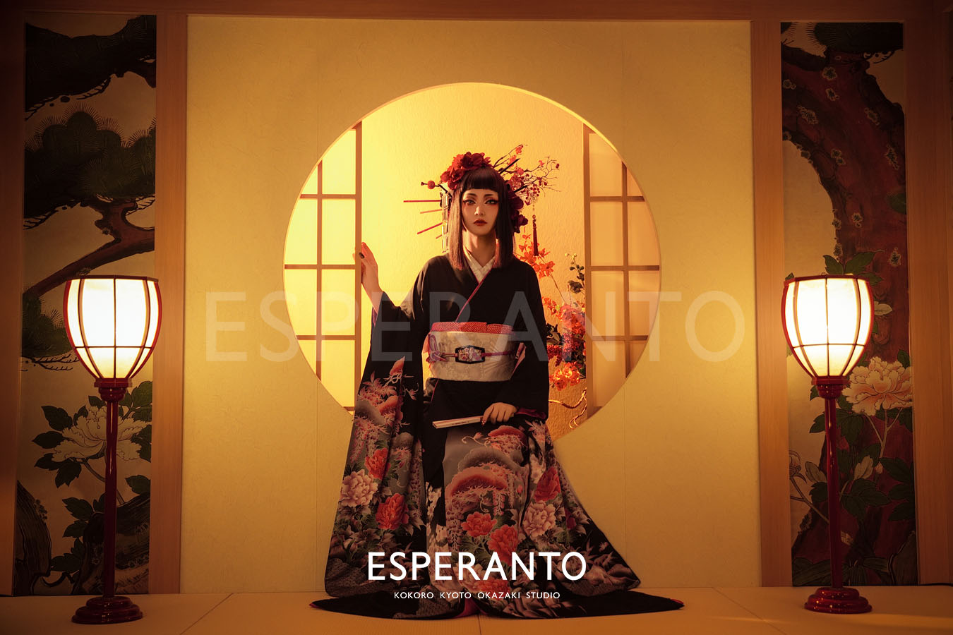 A guest in a black kimono with a peony pattern, before a gold moon window, lanterns on either side.