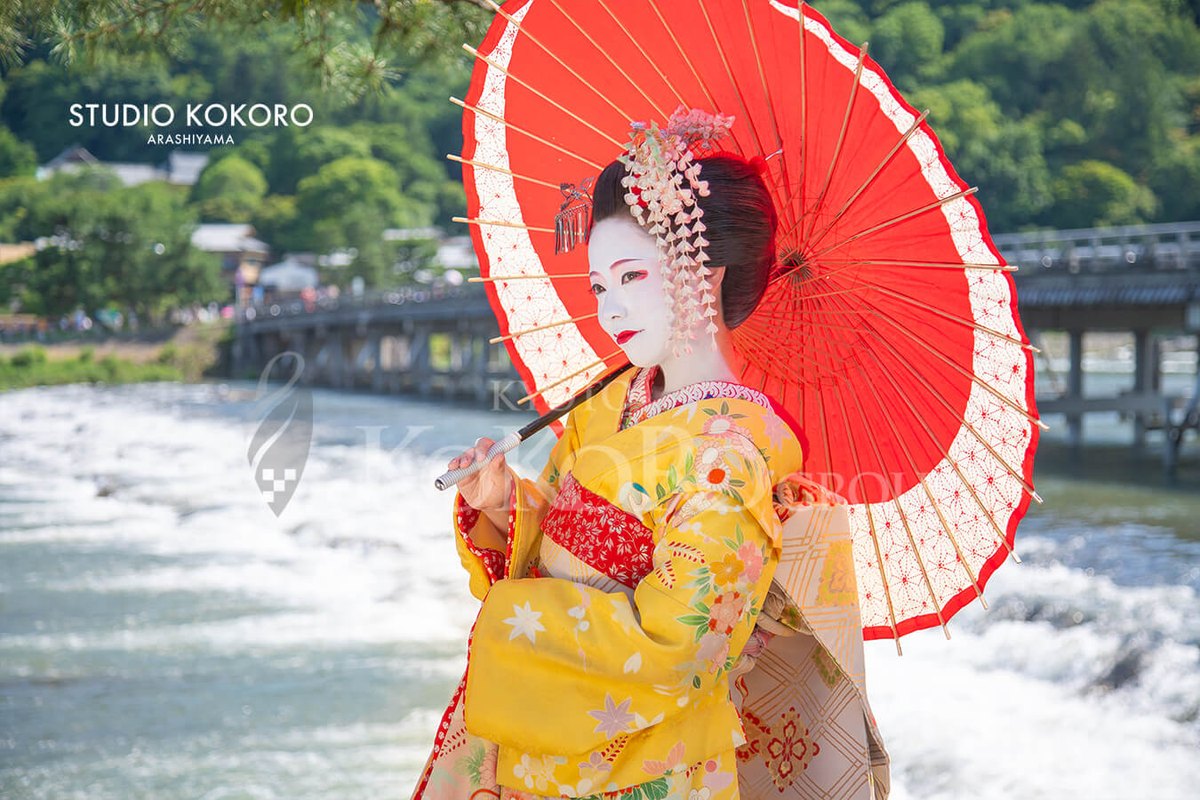 A guest dressed as a maiko, holding a red parasol, with Togetsukyo Bridge behind her.