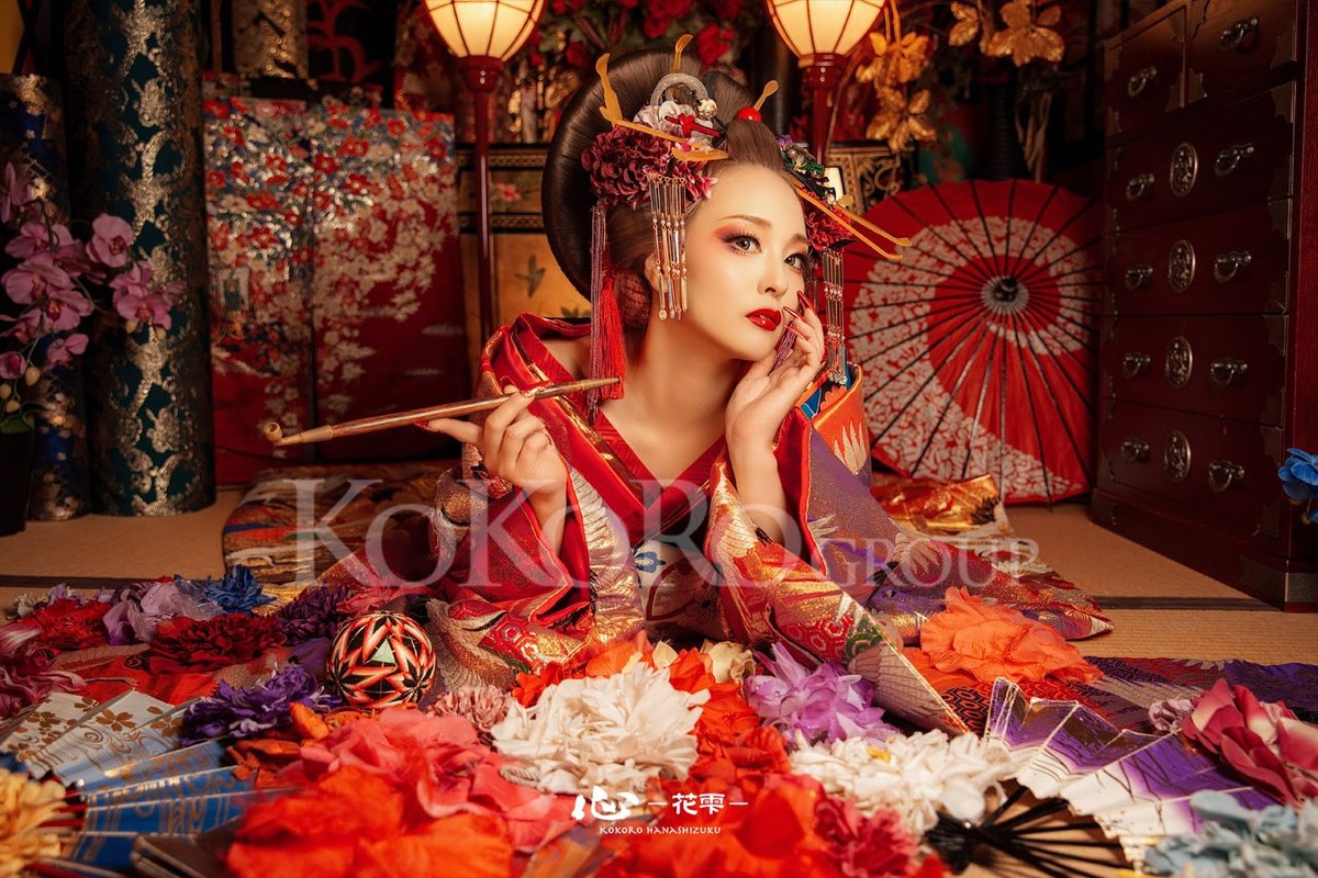 A guest dressed as an oiran, hairpins in place, on a tatami room strewn with fans and flowers.